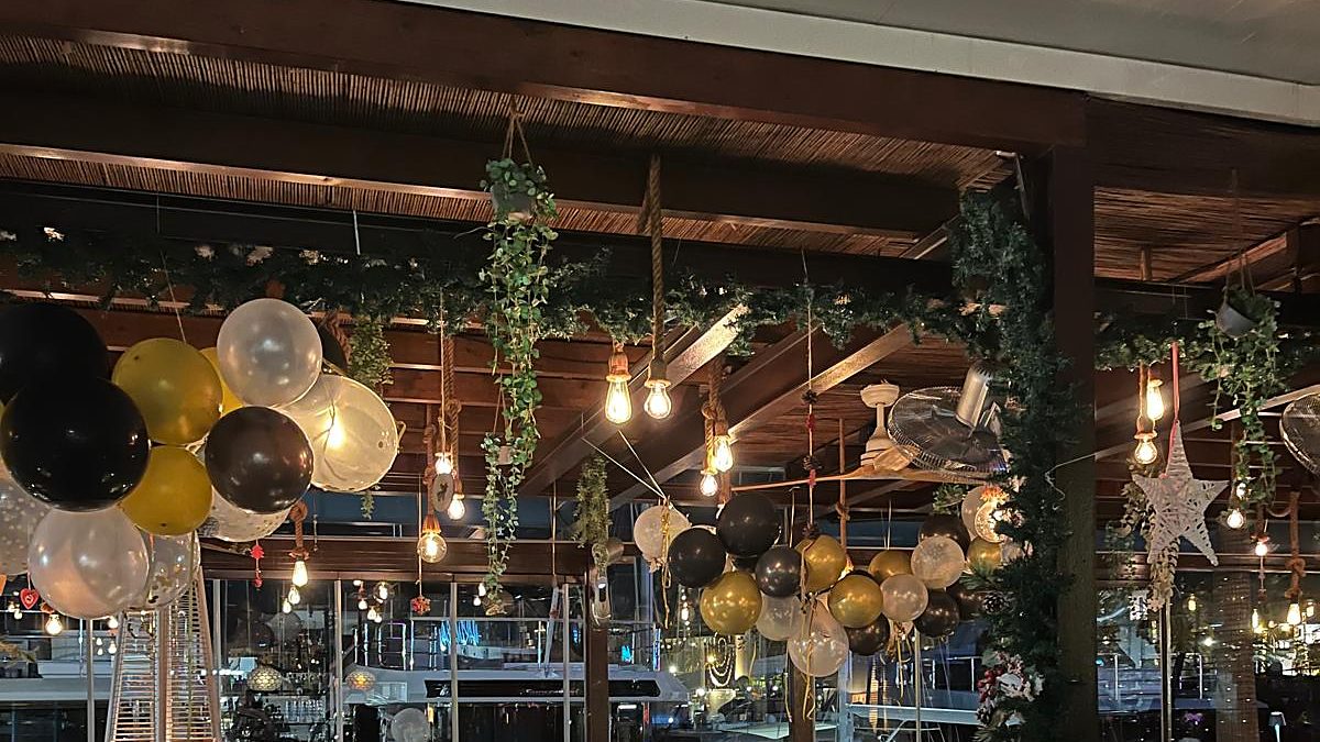 Techo del restaurante decorado con globos dorados y negros y bombillas, con los barcos del puerto de Palma al fondo